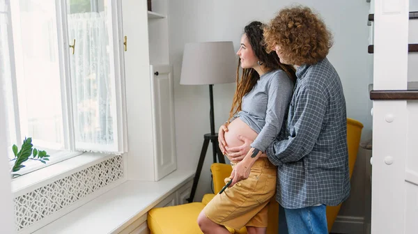 Glad hipster gravid par står tillsammans nära fönster i modern lägenhet och ömt röra magen. — Stockfoto