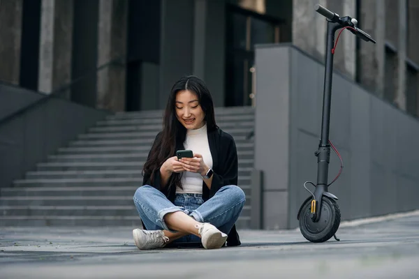 Uśmiechnięta, pewna siebie Azjatka siedzi na asfalcie w pobliżu skutera elektrycznego i używa smartfona. — Zdjęcie stockowe