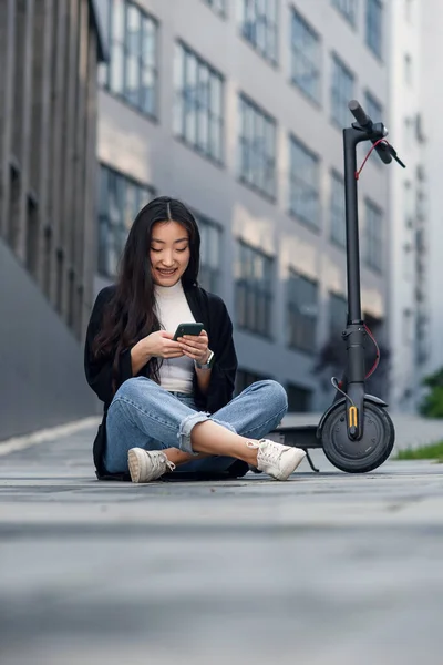 Улыбающаяся уверенная азиатка сидит на асфальте возле электроскутера и пользуется смартфоном. — стоковое фото