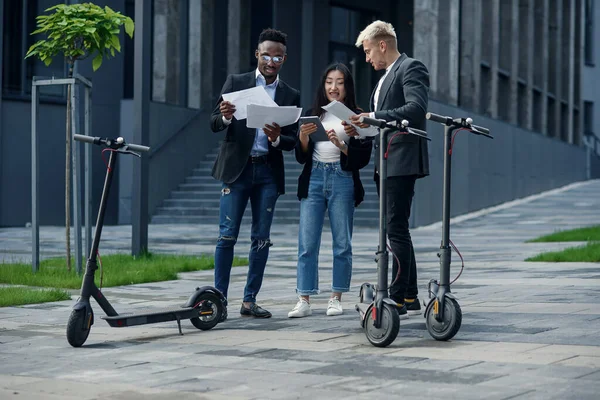 Equipo empresarial internacional responsable de tres empleados analiza proyecto conjunto cerca de edificio de oficinas cerca de e-scooters. — Foto de Stock