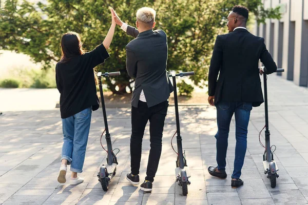 Vista trasera de alegres amigos internacionales sosteniendo scooters eléctricos que se acercan a un edificio moderno y hablan. — Foto de Stock