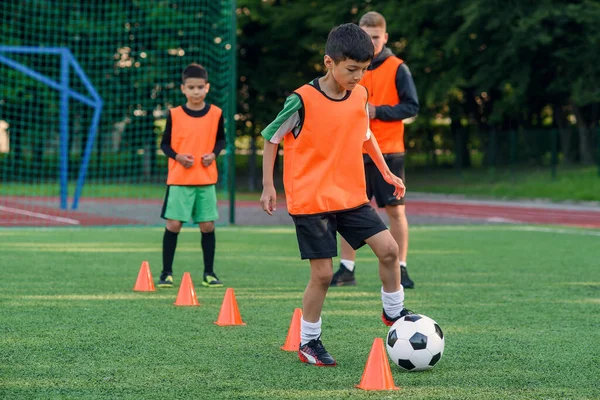 Přetrvávající dospívající chlapec ve sportovním oblečení vlaky fotbal na fotbalovém hřišti a učí se kroužit míč mezi tréninkovými kužely. — Stock fotografie