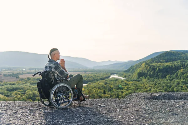 Можливо, недієздатний молодий хлопець сидить наодинці в інвалідному візку на пагорбі на красивому фоні природи і молиться . — стокове фото