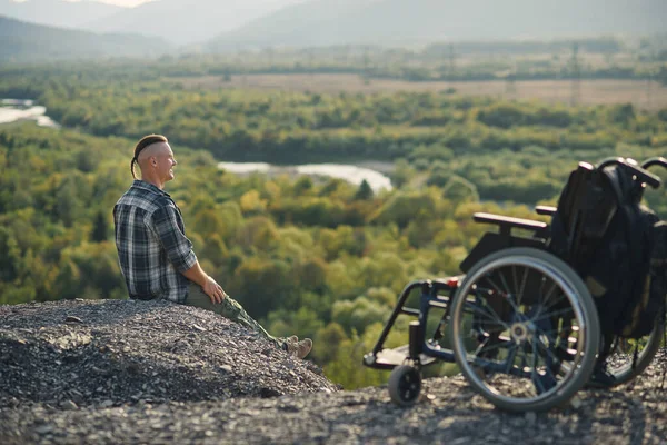 Молодий інвалід сидить на вершині гори біля інвалідного візка і насолоджується свіжим повітрям і красою природи. Насолоджуючись життям . — стокове фото