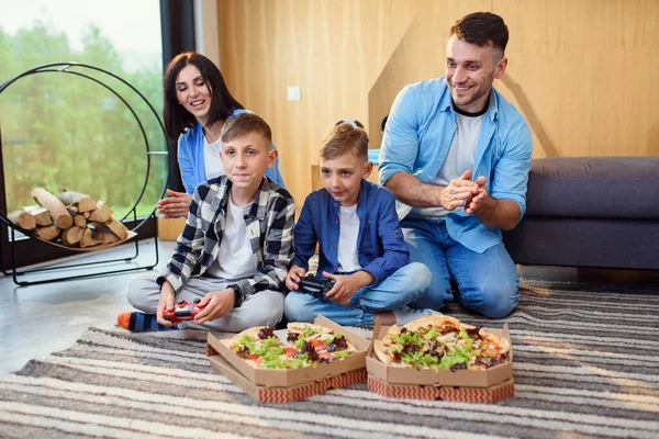 Pai feliz, mãe e dois filhos sentados no chão, jogando videogames com gamepads e comendo pizza saborosa. — Fotografia de Stock