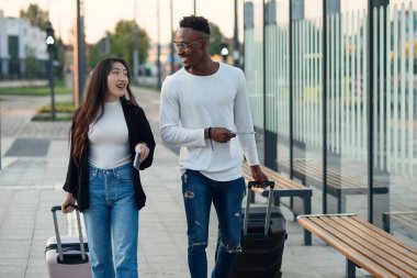 Pasaportları olan Afro-Amerikalı bir adam ve Asyalı bir kadın otobüs durağında bavullarını tutar ve konuşurlar..