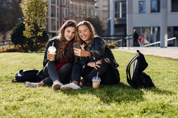 Duas elegantes meninas adolescentes atraentes com smoothie e smartphone sentado na grama. Tempo de lazer com os melhores amigos. — Fotografia de Stock