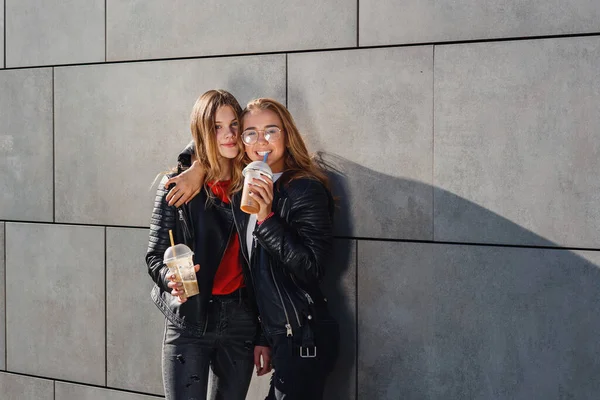 Duas namoradas adolescentes elegantes com smoothie posando perto de parede de concreto cinza. Tempo de lazer com os melhores amigos. — Fotografia de Stock