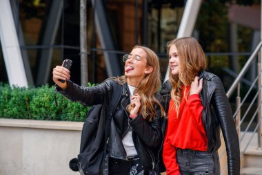 İki şık, mutlu, akıllı telefonlu genç kız caddedeki modern binanın yanında selfie çekiyorlar..
