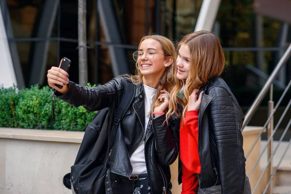 Duas meninas adolescentes felizes elegantes com smartphone fazendo selfie perto do elegante edifício moderno na rua. — Fotografia de Stock