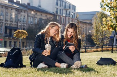 Smoothie ve akıllı telefonlu iki çekici genç kız çimlerin üzerinde oturuyor. En iyi arkadaşlarla boş vakit geçirmek.