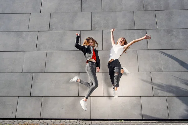 İki güzel genç kız arkadaş atlayıp gri beton duvarın yanında eğleniyorlar. En iyi arkadaşlarla boş vakit geçirmek. — Stok fotoğraf