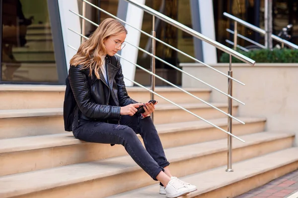 Veselá krásná studentka používá chytrý telefon, zatímco sedí na schodech na ulici, těší procházka venku. — Stock fotografie