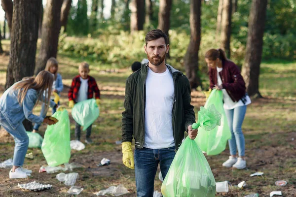 Handsome bearded man with full rubbish pack in background of park where his friends activists picking up rubbish.