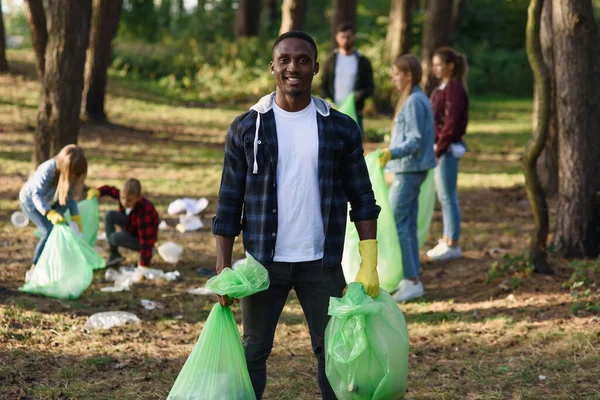 Stylish bearded black man with full rubbish packs on background of his friends activists picking up rubbish at forest.