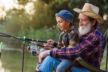 Likable respected 70-aged bearded grandfather holding on knees 10-aged cute grandson and teaching him to catch fish. clipart
