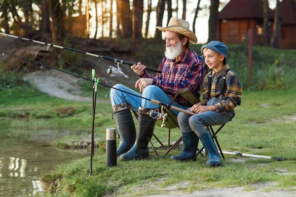 Happy boy sitting on chairs together with his experienced old grey-bearded granddad and fishing with rods on the lake. — Stock Photo, Image