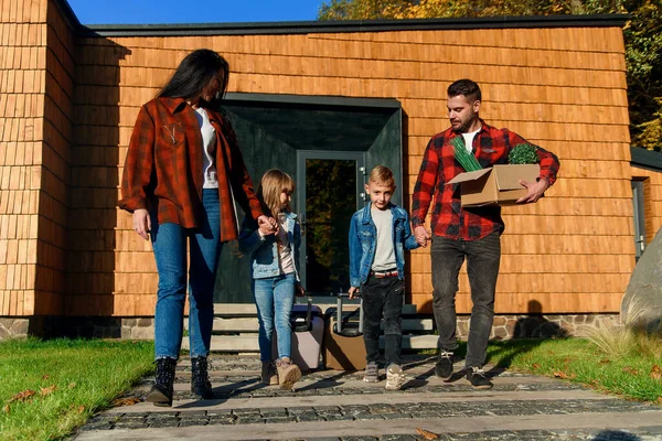 Högpiga barn och deras lyckliga föräldrar går tillsammans till sin bil med resväskor och kartong med gröna blommor. — Stockfoto