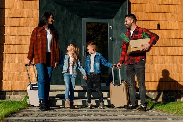 Högpiga barn och deras lyckliga föräldrar går tillsammans till sin bil med resväskor och kartong med gröna blommor. — Stockfoto