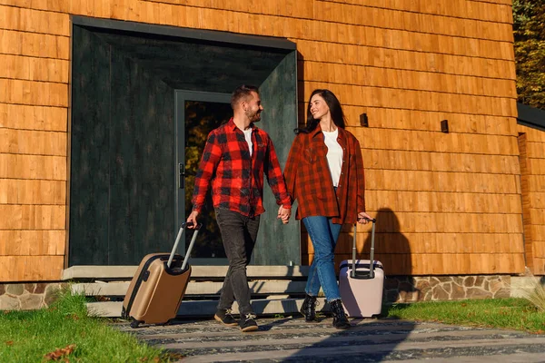 Homem barbudo bonito e mulher muito elegante saindo da casa moderna com malas e indo juntos para tropeçar. — Fotografia de Stock