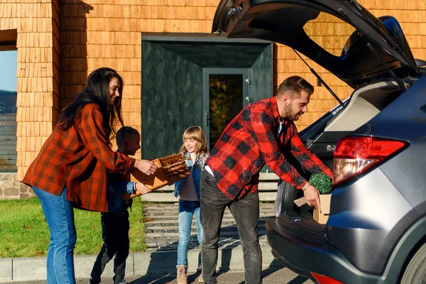 Família alegre ajudando uns aos outros para descarregar o tronco de automóveis de caixas de papelão e malas durante a mudança para novo apartamento — Fotografia de Stock