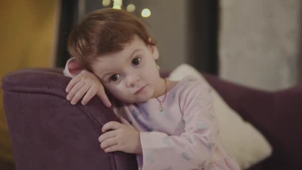 Little girl is sitting on the couch and is looking at the camera — Stock Video