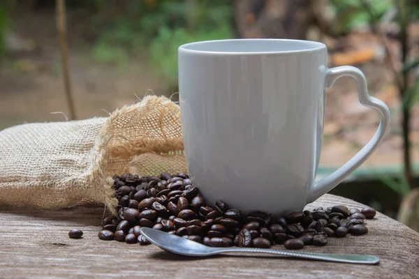 Cup of coffee with smoke and coffee beans in burlap sack on wooden table and blurred tree background.