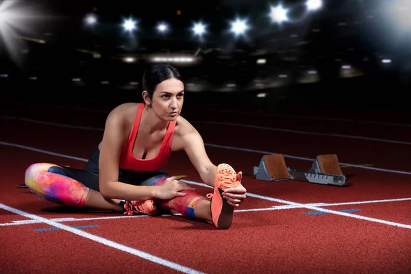 Ung atletisk kvinna gör övningar på flexibilitet. Stretching benen sitter spår stadion, natt med reflektorer — Stockfoto