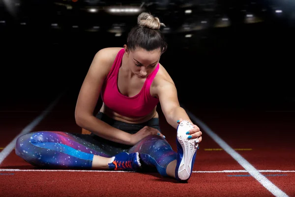 Young athletic woman doing exercises on flexibility. stretching legs sitting track stadium, night with reflectors — Stock Photo, Image