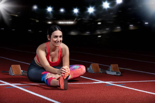 Ung atletisk kvinna gör övningar på flexibilitet. Stretching benen sitter spår stadion, natt med reflektorer — Stockfoto