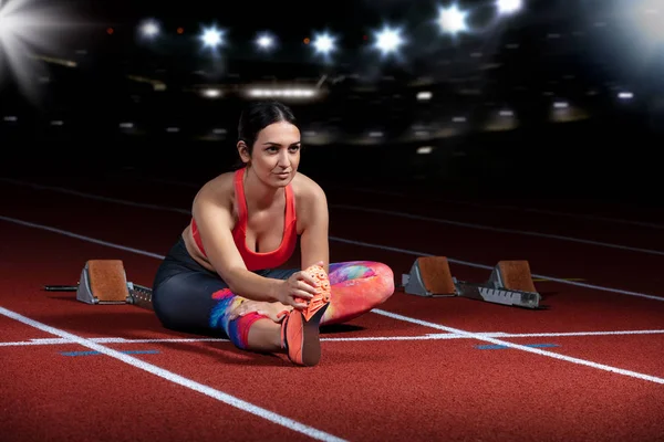 Ung atletisk kvinna gör övningar på flexibilitet. Stretching benen sitter spår stadion, natt med reflektorer — Stockfoto