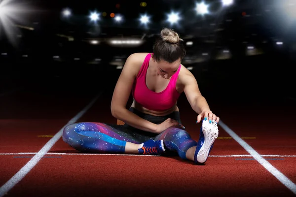 Young athletic woman doing exercises on flexibility. stretching legs sitting track stadium, night with reflectors — Stock Photo, Image