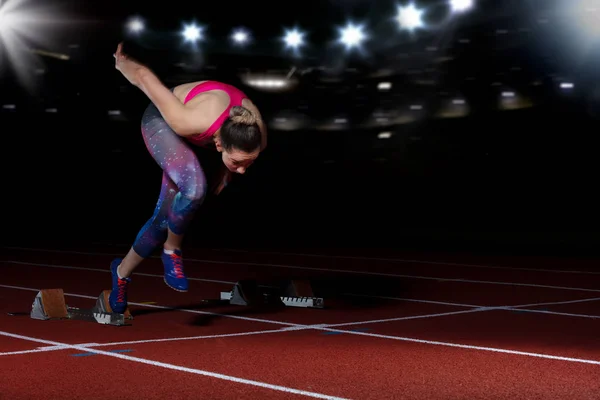 Женщина-спринтер покидает стартовые блоки на атлетическом треке. Взрывной старт на стадионе с отражателями — стоковое фото