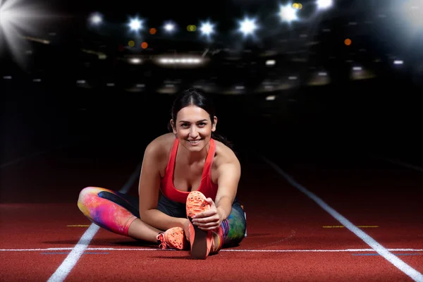 Mujer atlética joven haciendo ejercicios de flexibilidad. estiramiento piernas sentado pista estadio, noche con reflectores — Foto de Stock