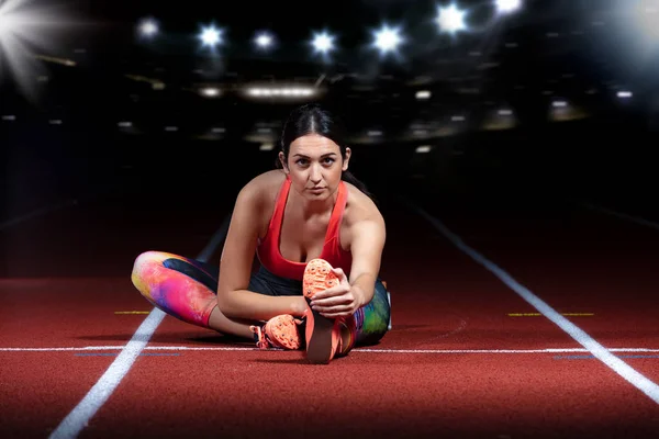 Young athletic woman doing exercises on flexibility. stretching legs sitting track stadium, night with reflectors — Stock Photo, Image