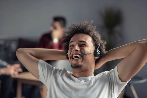 Zadowolony zrelaksowany Afro-freelancer, czuje się szczęśliwy w pracy siedzi biurko z zestawu słuchawkowego. — Zdjęcie stockowe