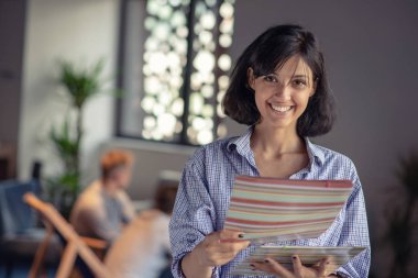Akıllı rahat kıyafet güzel Afro Amerikan iş kadını kameraya bakarak ve ofiste bir klasörle dururken gülümseyen.