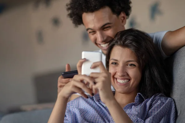 Casal atraente sentado no sofá juntos olhando para o smartphone em casa na sala de estar — Fotografia de Stock