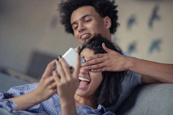 Atractiva pareja sentada en el sofá juntos mirando el teléfono inteligente en casa en la sala de estar — Foto de Stock