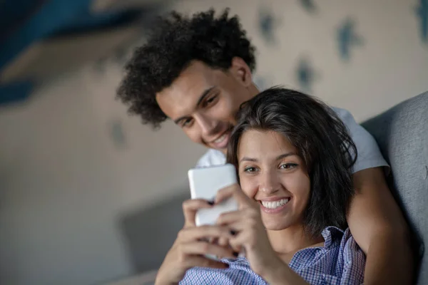 Atractiva pareja sentada en el sofá juntos mirando el teléfono inteligente en casa en la sala de estar — Foto de Stock