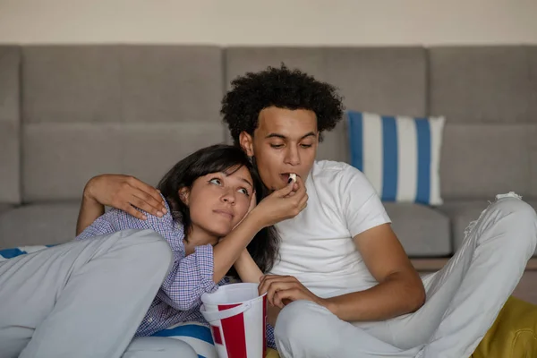 Imagem de casal sorridente sentado no chão perto do sofá em casa e olhando para o lado enquanto come pipocas — Fotografia de Stock