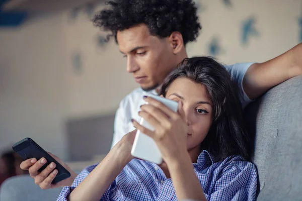 Casal atraente sentado no sofá juntos olhando para o smartphone em casa na sala de estar — Fotografia de Stock