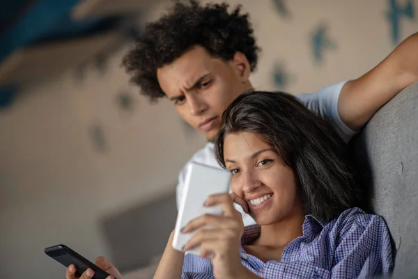 Atractiva pareja sentada en el sofá juntos mirando el teléfono inteligente en casa en la sala de estar — Foto de Stock