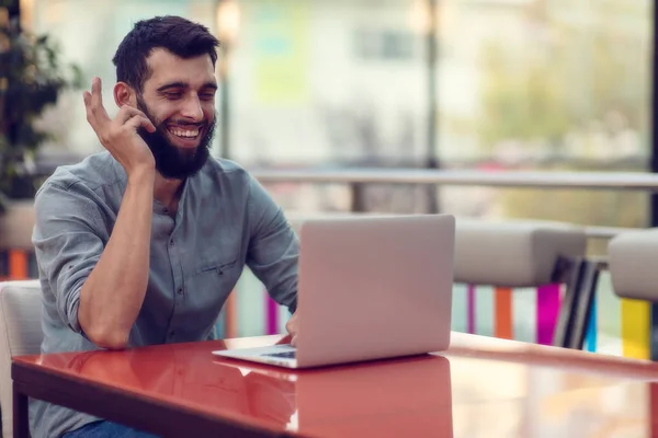 Retrato de meio comprimento de designer barbudo bem sucedido sorrindo para a câmera enquanto trabalhava em freelance no netbook . — Fotografia de Stock