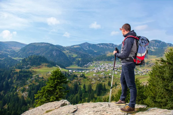 Hiker young man with backpack and trekking poles standing on edge of cliff and looking at the lake, rear view — Stock Photo, Image