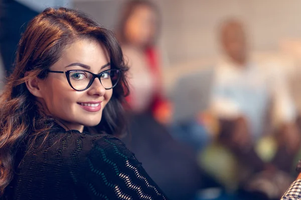 Jovem empresária usando óculos sorrindo confiantemente enquanto estava em um escritório com colegas trabalhando em segundo plano — Fotografia de Stock