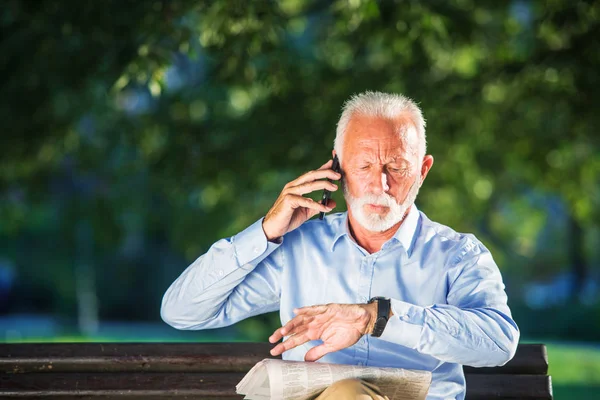 Sonriente hombre mayor hablando por teléfono mientras está sentado en el banco en el parque . — Foto de Stock