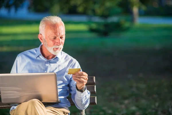 Vecchio maschio seduto sulla panchina e surf net sul computer portatile nel parco, shopping on-line concetto — Foto Stock