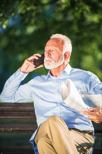 Sonriente hombre mayor hablando por teléfono mientras está sentado en el banco en el parque . — Foto de Stock
