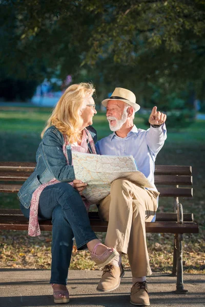 Retrato de hombre y mujer felices leyendo mapa mientras están sentados en un banco del parque. Pareja mayor de vacaciones usando el mapa de la ciudad . — Foto de Stock